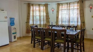 A dining area with a wooden table and chairs, a water dispenser, and curtains on the windows. - Imanisuites (Nairobi)