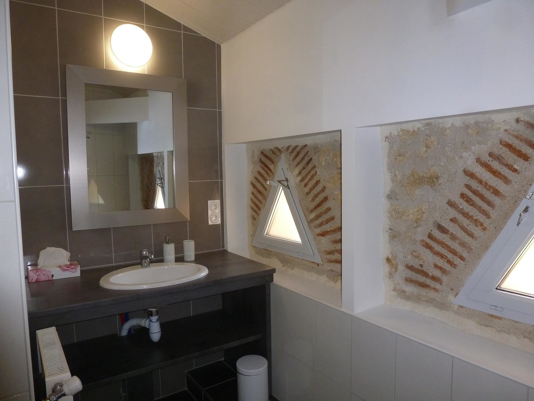 A bathroom with a large mirror, a sink, and a shower. There are two windows with triangular frames and a textured wall.