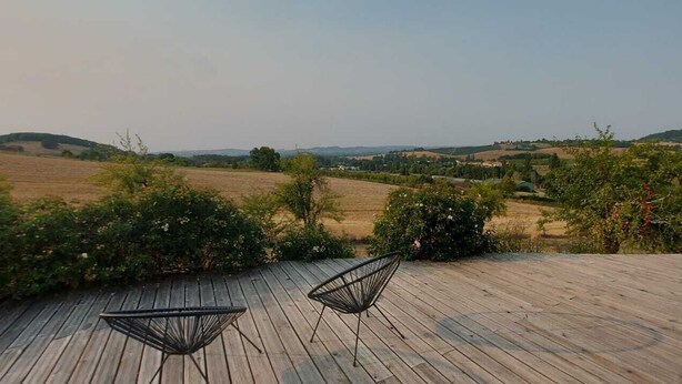 Une terrasse en bois avec deux chaises en métal, donnant sur un paysage rural composé de champs et de collines.