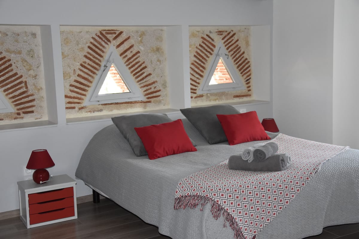 Une chambre à coucher moderne avec un lit, des coussins rouges et gris, un plaid à motifs, une table de chevet avec une lampe rouge et une petite commode.