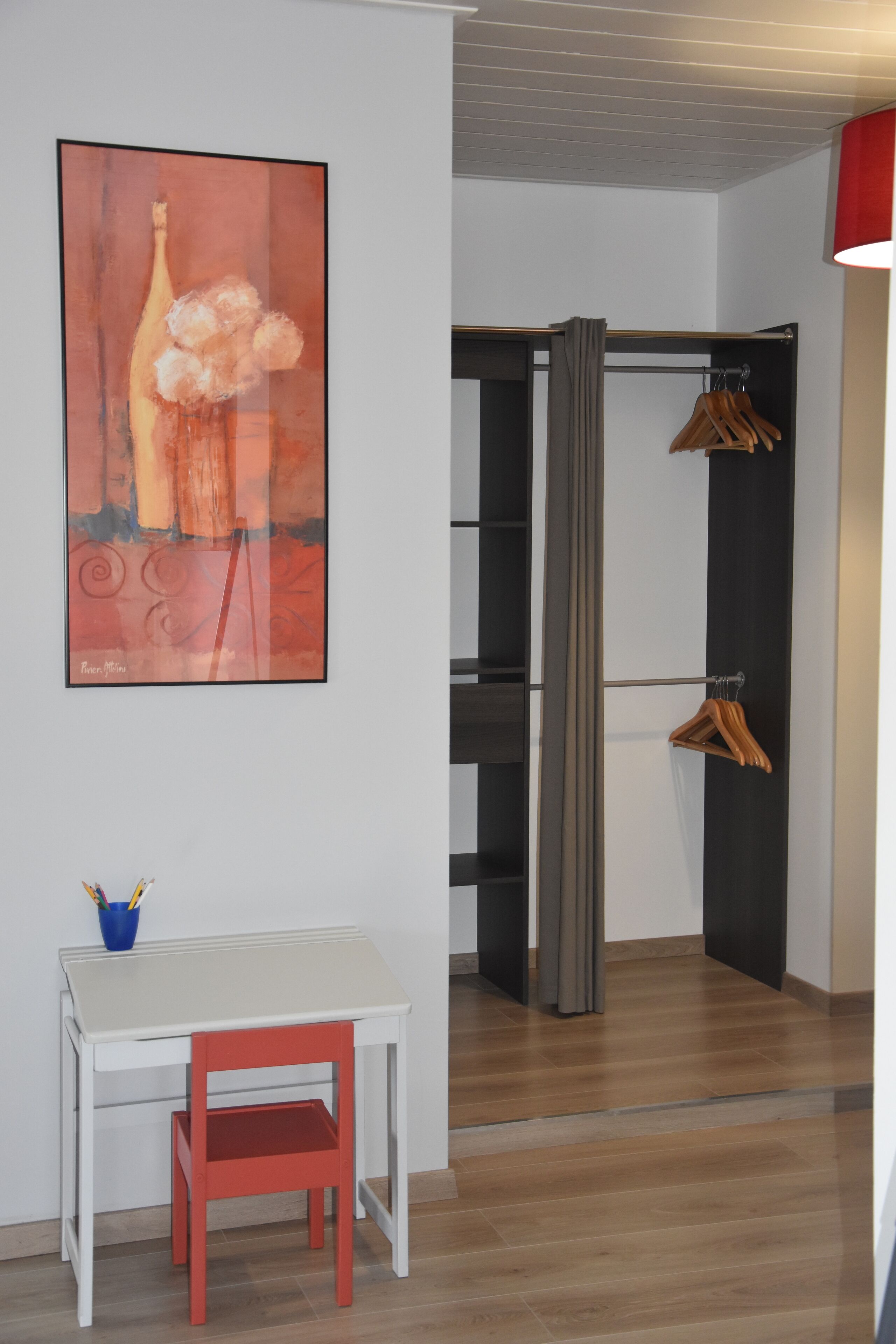 Una habitación con un escritorio blanco, una silla roja, un cuadro abstracto grande, un armario con ropa colgada y una lámpara roja.