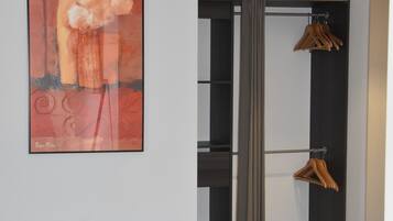 Una habitación con un escritorio blanco, una silla roja, un cuadro abstracto grande, un armario con ropa colgada y una lámpara roja.