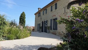 Un bâtiment en pierre de deux étages, avec une allée gravillonnée et un jardin agrémenté de fleurs violettes.