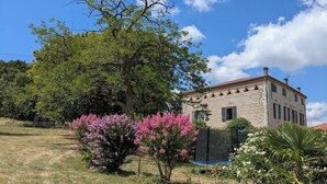 Un bâtiment en pierre, surmonté d’un toit en tuiles, entouré de pelouse et d’arbustes en fleurs.