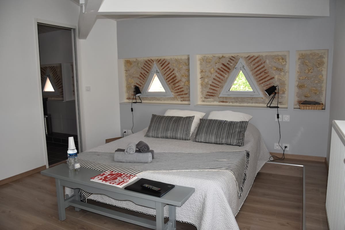 Une chambre à coucher avec un lit, des lampes de chevet, une table avec un livre et une fenêtre donnant sur un mur de briques.