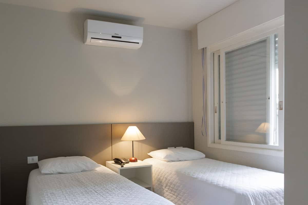 A hotel room with two single beds, a bedside table with a lamp, an air conditioning unit, and a window with blinds.
