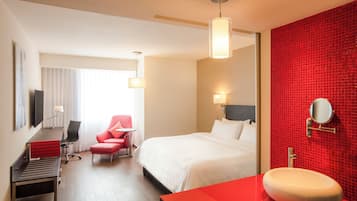 Una habitación de hotel con cama, escritorio, silla, una pared de acento roja, una mesita de noche roja y un baño con pared de azulejos rojos y un espejo redondo.