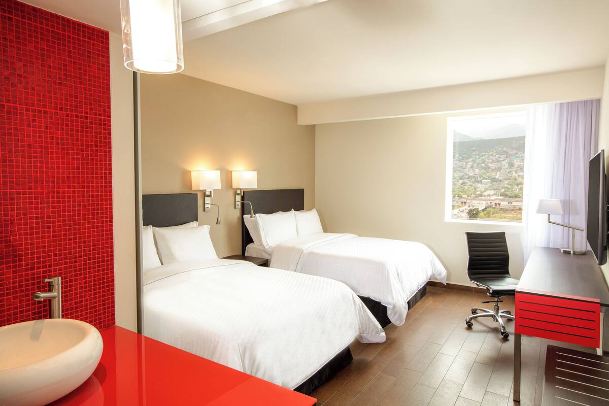 A hotel room with two beds, a desk, a chair, a TV, and a bathroom with a red-tiled wall.