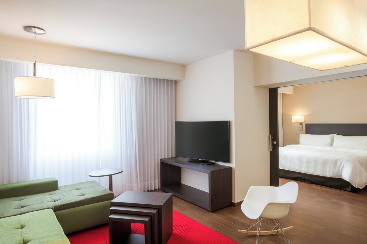A hotel room with a bed, a TV on a stand, a green sofa, a small table, and a white chair.