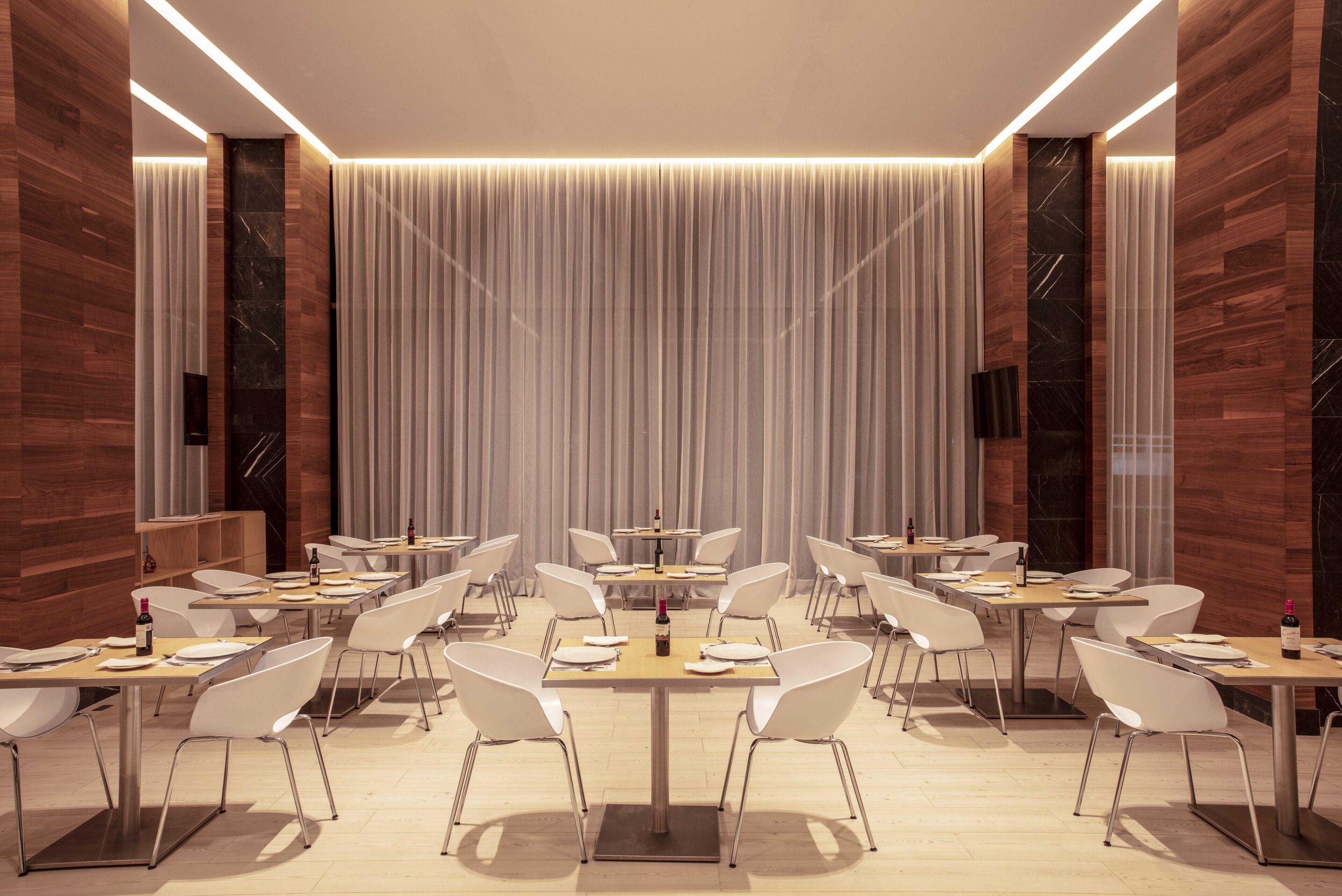A modern dining area with multiple tables and white chairs, featuring a central column and a backdrop of vertical drapery.