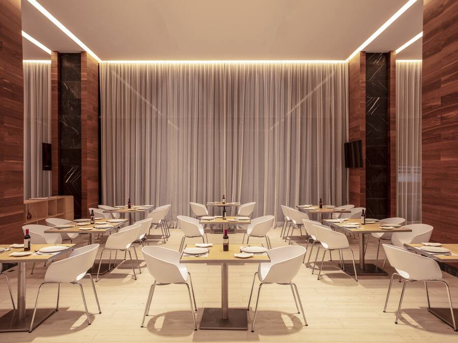 A modern dining area with multiple tables and white chairs, featuring a central column and a backdrop of vertical drapery.