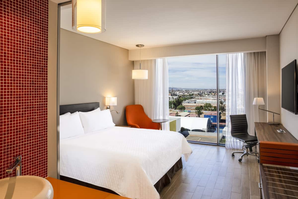 A hotel room with a bed, a desk, a chair, a large window with a city view, and a red-tiled wall.