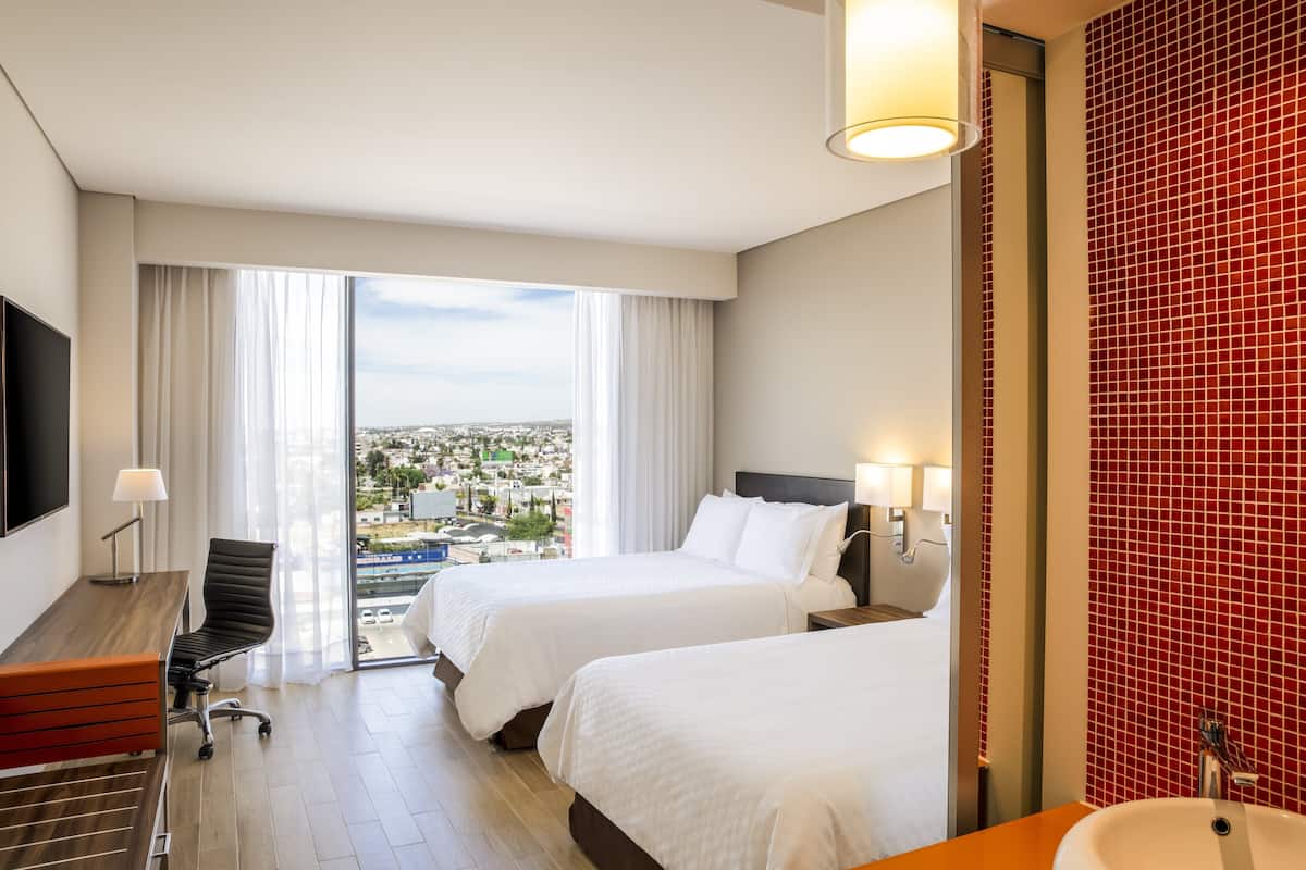 A hotel room with two beds, a desk, a chair, a large window with a city view, and a red-tiled wall.