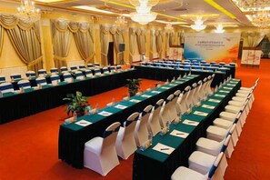 Un salón de conferencias con mesas y sillas dispuestas en filas, un escenario y un cartel.