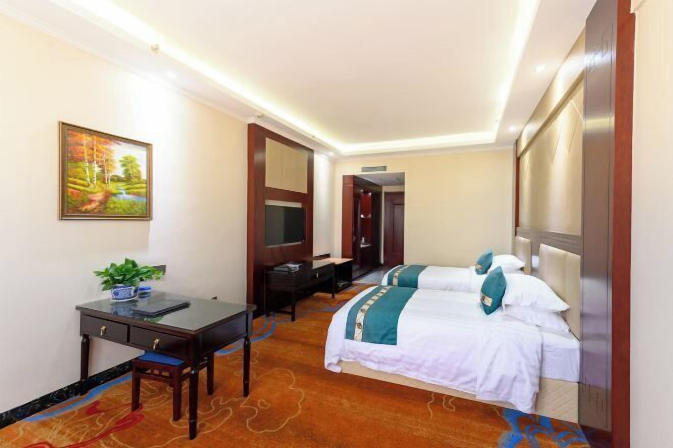 A hotel room with two beds, a desk with a chair, a small table with a plant, and a painting on the wall.