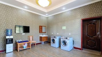 A hotel room with a television, a washing machine, a sink, and a wooden bench.