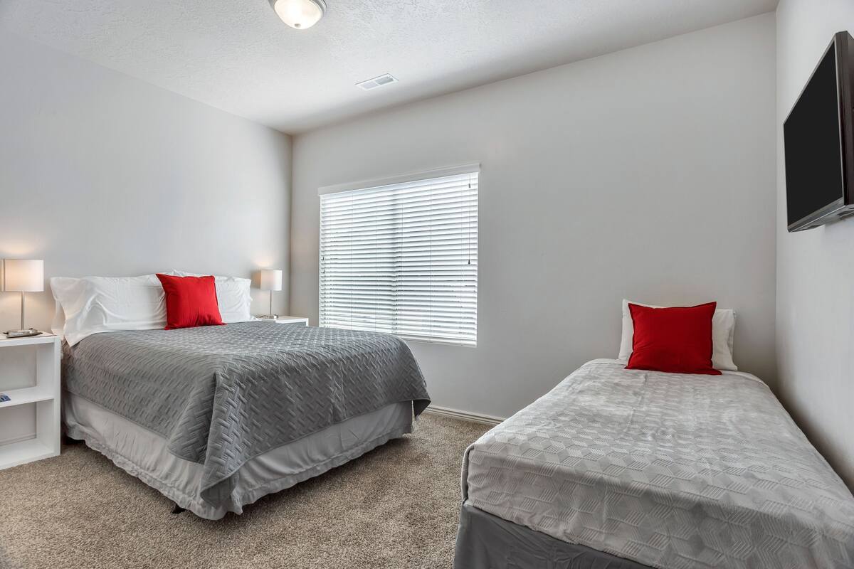 A bedroom with two beds, each with a red and white pillow, and a window with blinds.