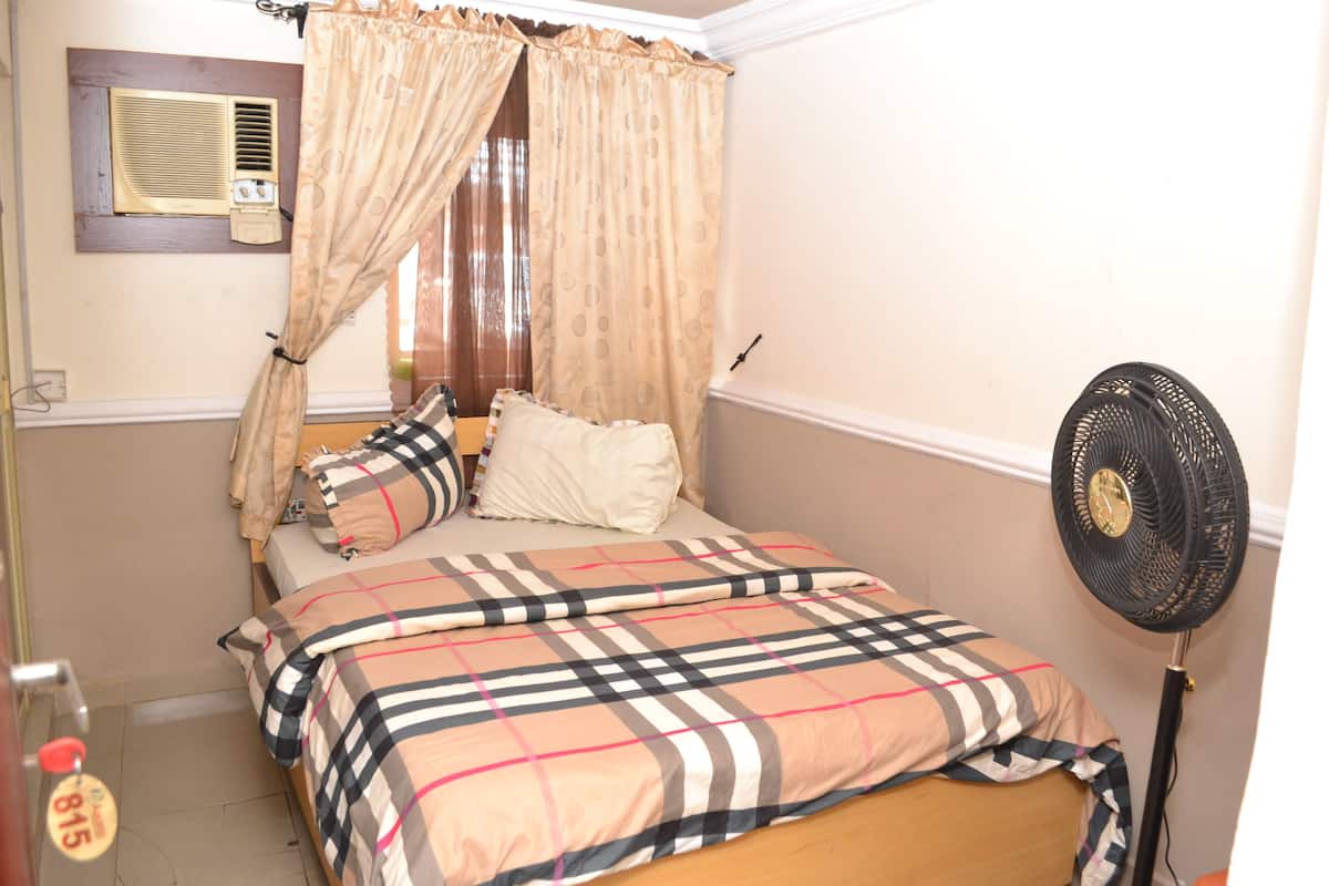 A bedroom with a bed, curtains, a standing fan, and a window with an air conditioning unit.