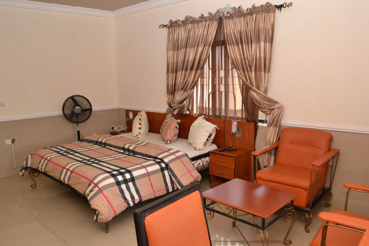 A hotel room with a bed, a chair, a table, a fan, and curtains.