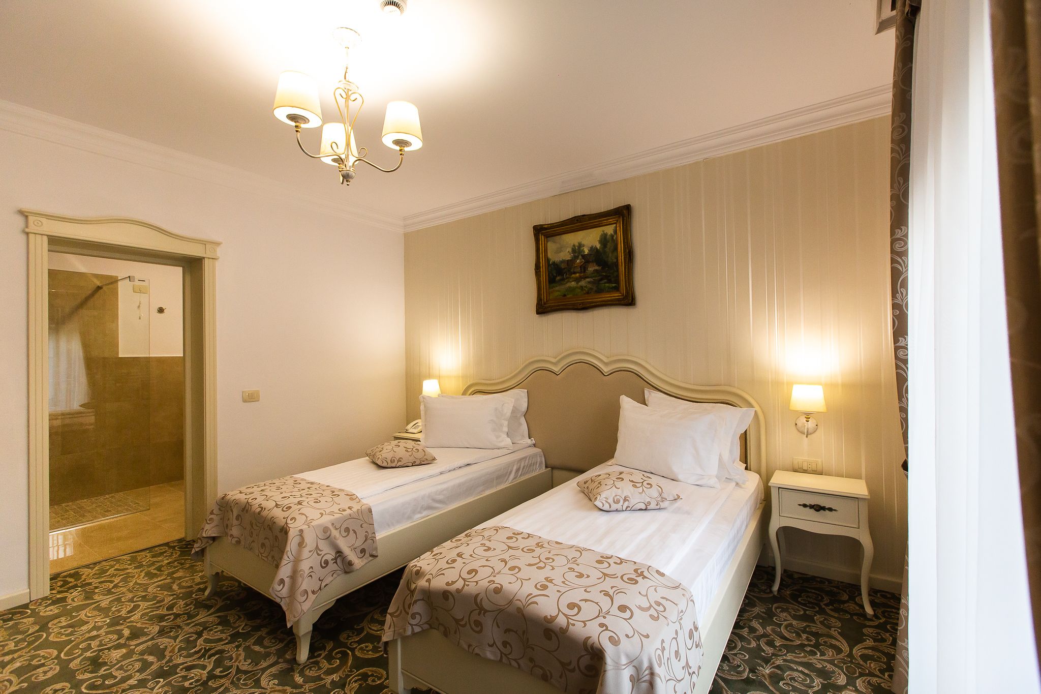 A hotel room with two beds, a chandelier, a painting on the wall, and a door leading to a bathroom.
