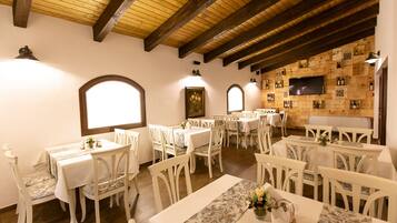 Un espace repas avec des tables et des chaises blanches, des poutres en bois au plafond et un mur décoré de bouteilles de vin.
