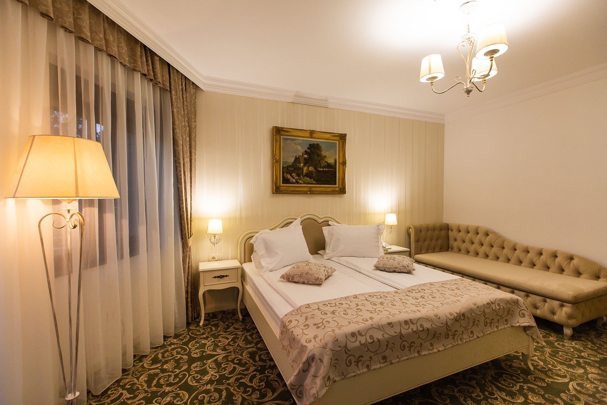 A hotel room with a bed, a sofa, a nightstand, a lamp, and a painting on the wall.