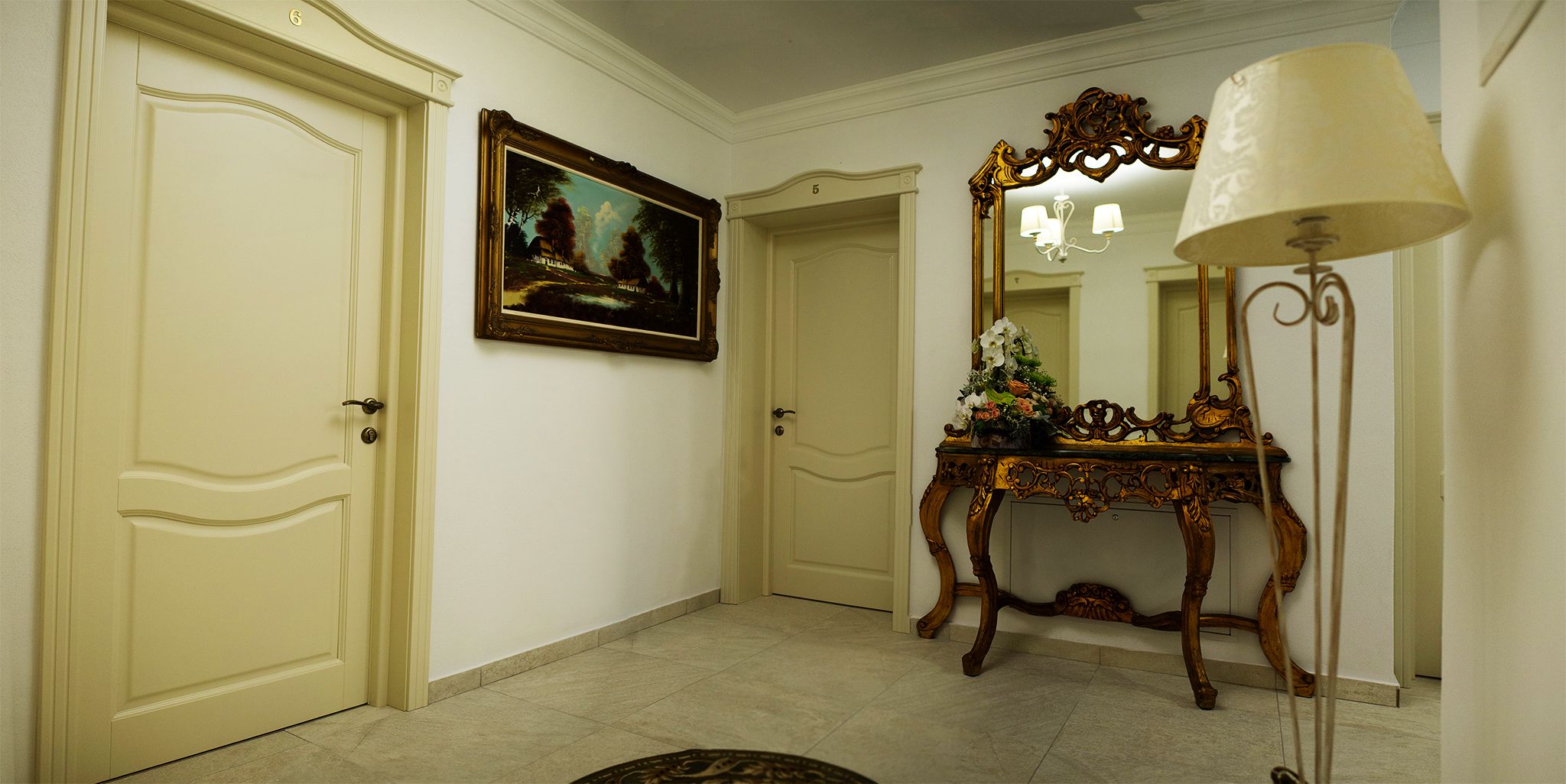 A hallway with cream-colored doors, a framed landscape painting, a mirror with an ornate frame, and a table with a floral arrangement.