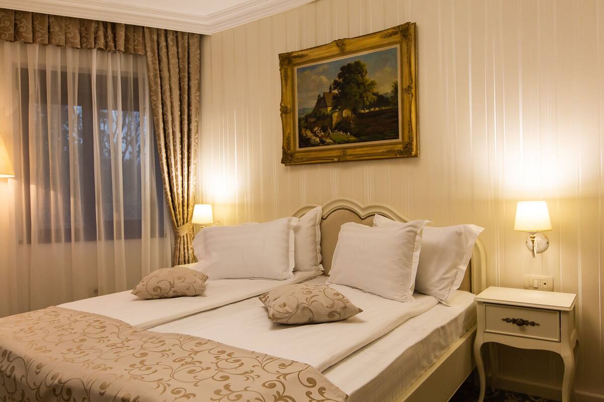 A double bed with a patterned bedspread and two pillows, a nightstand with a lamp, and a framed painting on the wall.
