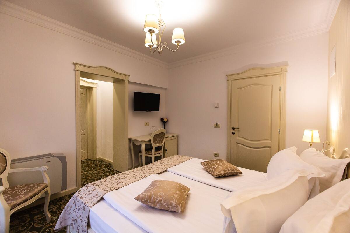 A hotel room with two beds, a desk, a chair, a television, and a chandelier.