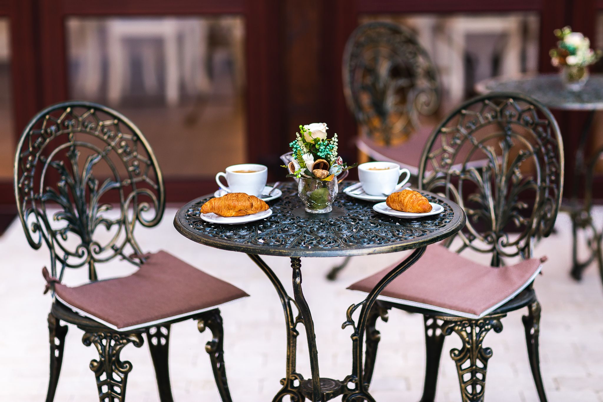 A round table with two chairs, each with a cup of coffee and a plate of croissants. A vase with flowers is on the table.