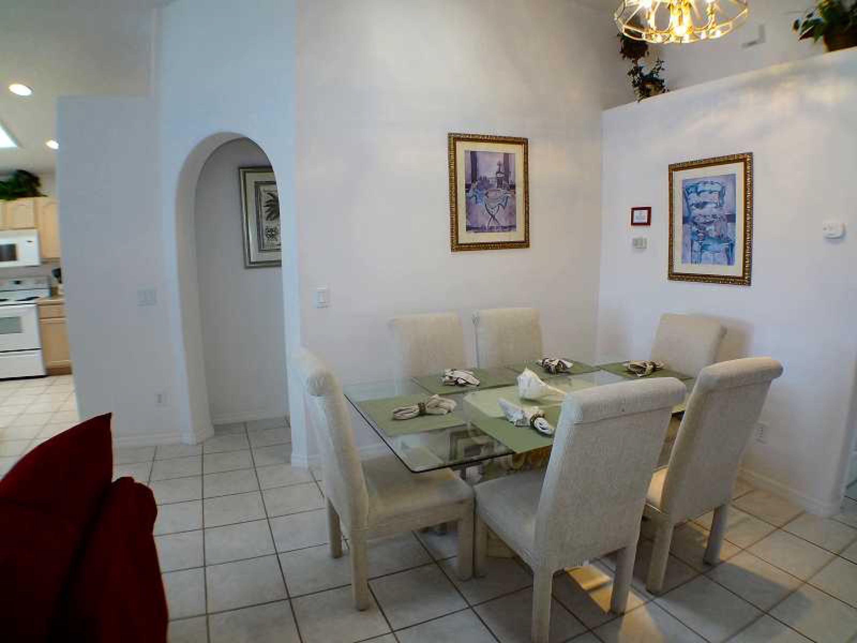 A dining area with a glass table and four chairs, a kitchen in the background, and two framed pictures on the wall.