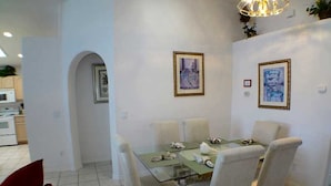A dining area with a glass table and four chairs, a kitchen in the background, and two framed pictures on the wall.