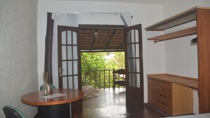 A room with a wooden desk, a chair, a small table, and a balcony with a view of greenery. - Hospedagem Praia de Piratininga (Niteroi)