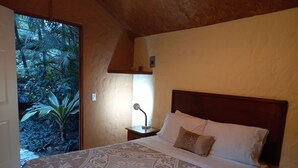 A bedroom with a bed, a nightstand, a lamp, and a view of a tropical garden.