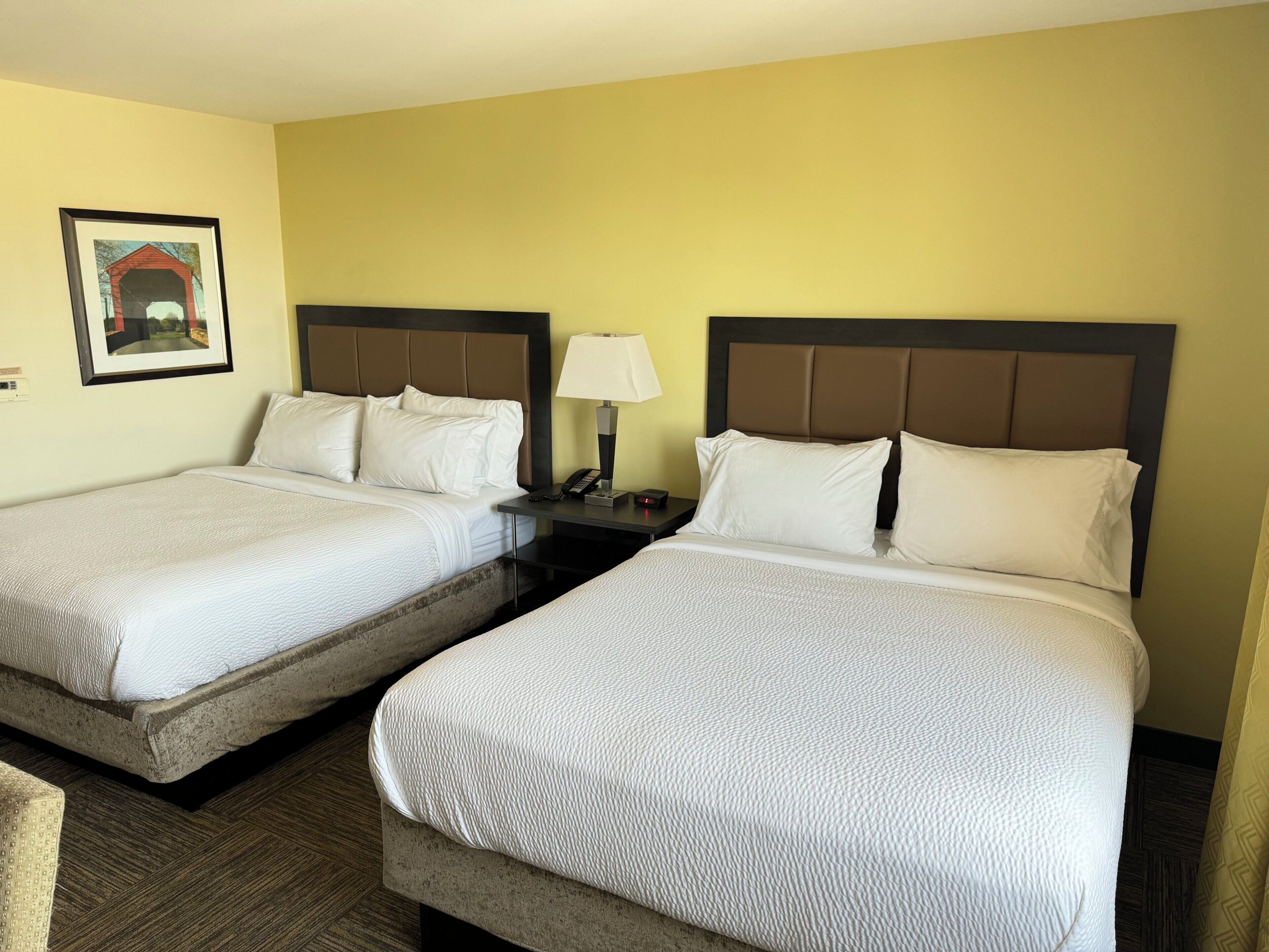 A hotel room with two beds, each with a headboard, a bedside table with a lamp, and a framed picture on the wall.