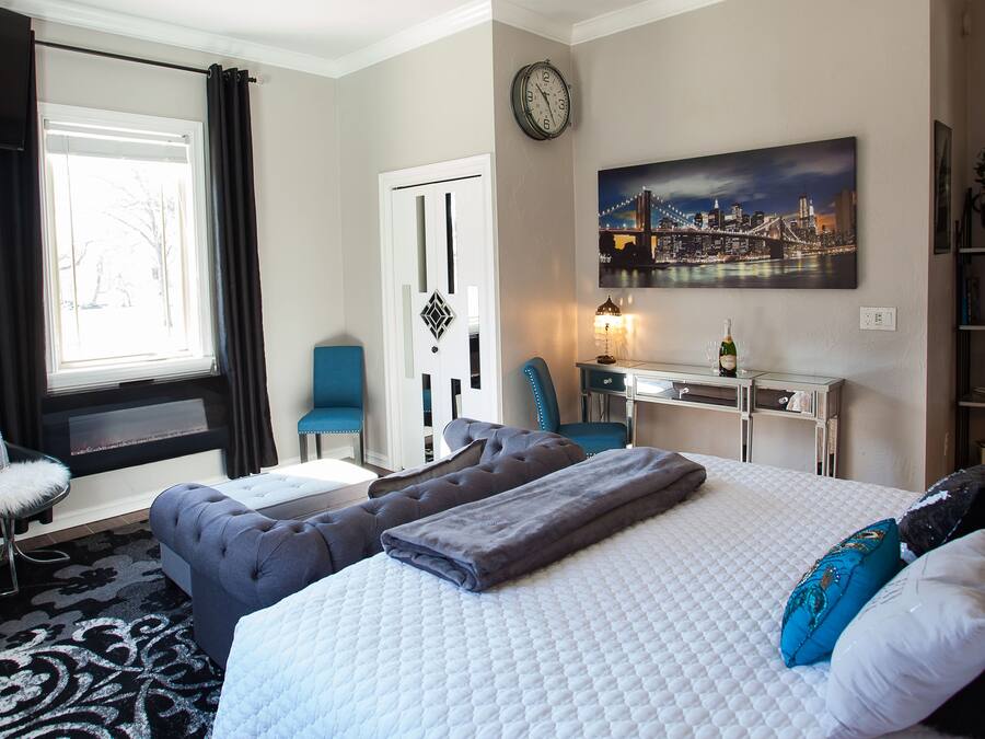A hotel room with a bed, a desk, a chair, a sofa, a fireplace, and a cityscape painting on the wall.