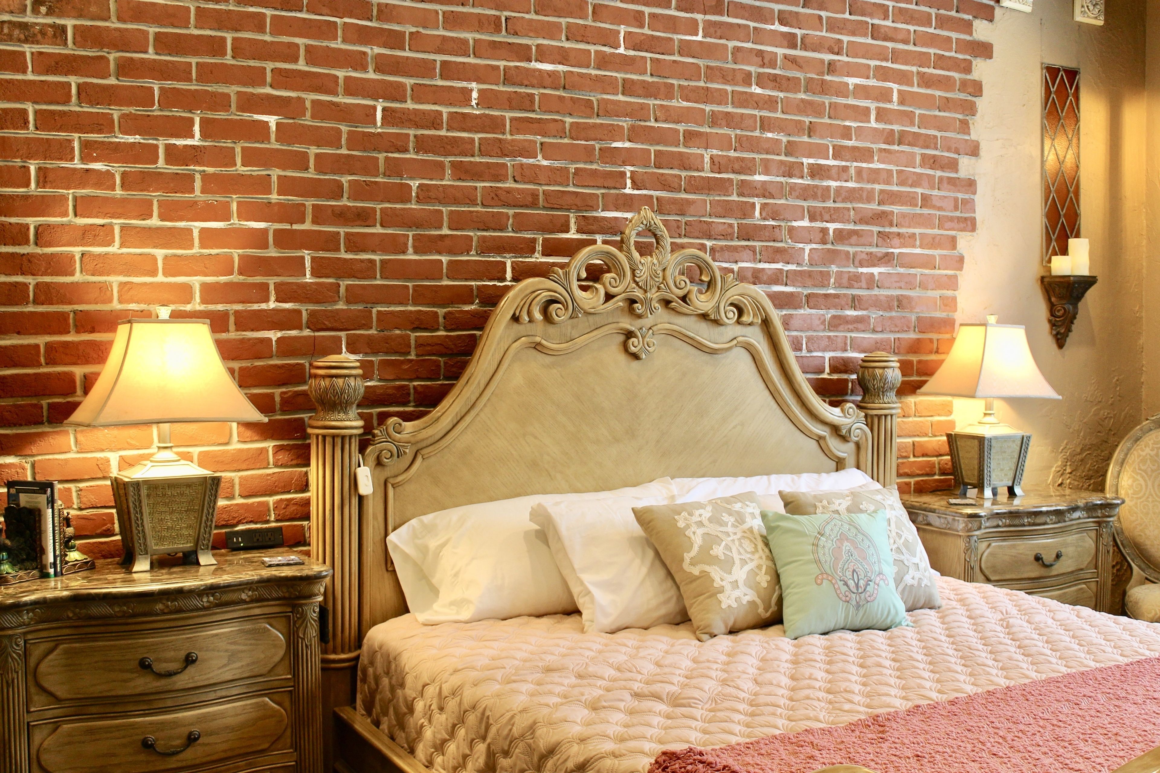 Un dormitorio con pared de ladrillo, cabecera de madera, dos mesitas de noche con lámparas y una cama con muchos almohadones.