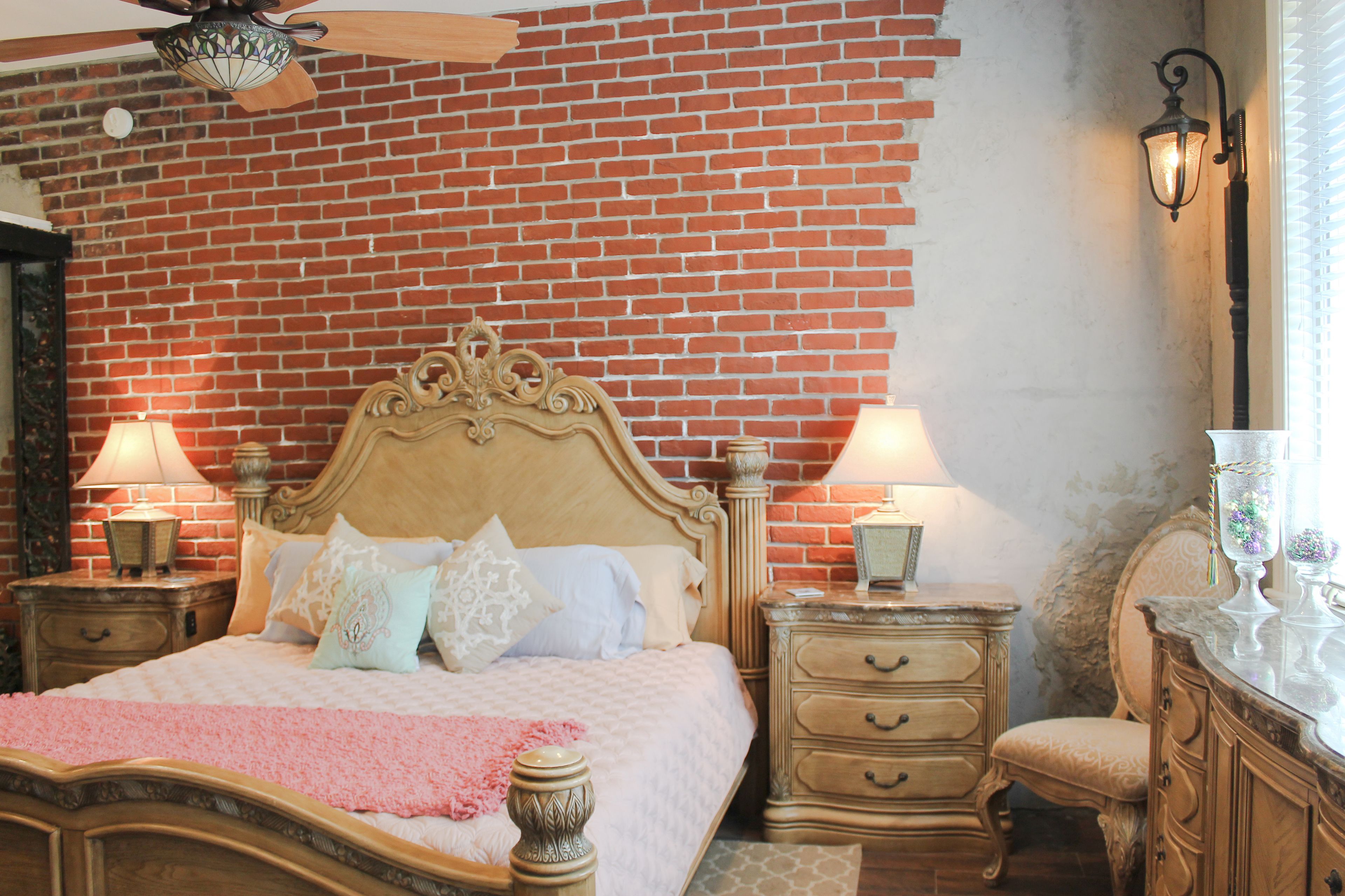 A bedroom with a brick wall, a wooden bed with ornate headboard, two bedside tables with lamps, a nightstand, a dresser, and a chair.