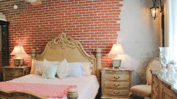 A bedroom with a brick wall, a wooden bed with ornate headboard, two bedside tables with lamps, a nightstand, a dresser, and a chair.