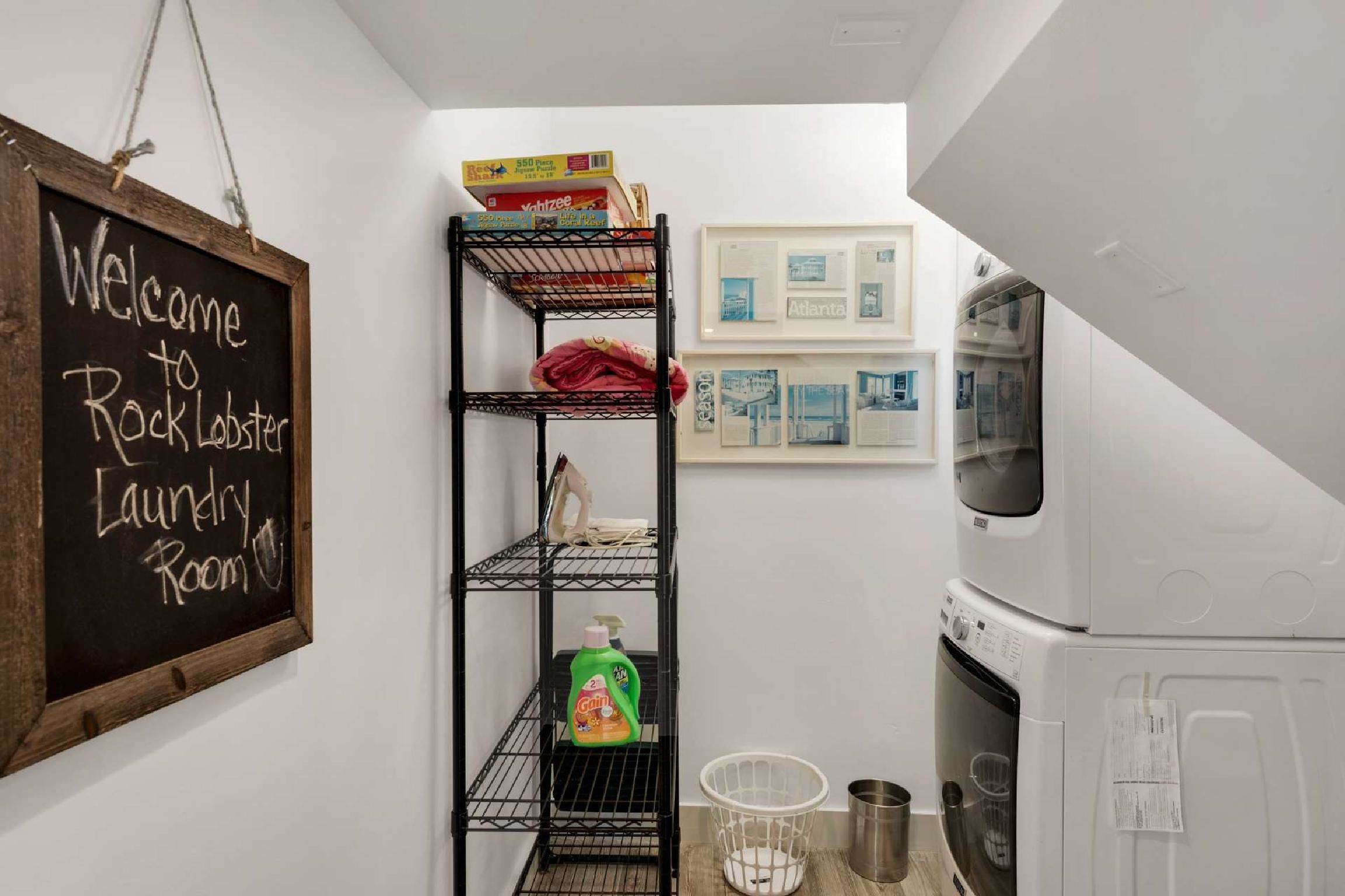 A laundry room with a chalkboard sign, a metal shelving unit with various items, a washing machine, and framed pictures on the wall.