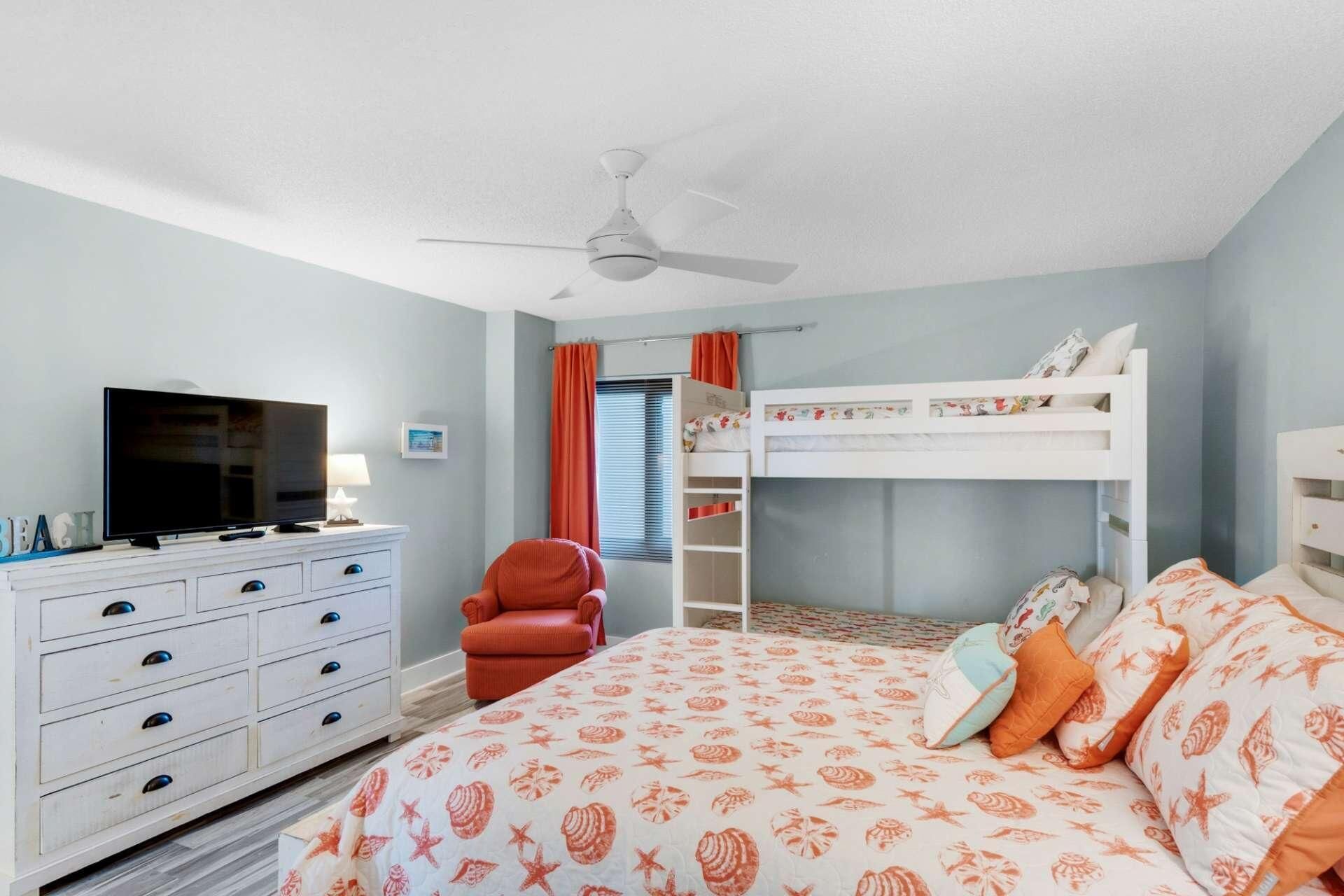 Ein Schlafzimmer mit Etagenbett, Fernseher auf einer Kommode, einem Sessel und einem Deckenventilator.