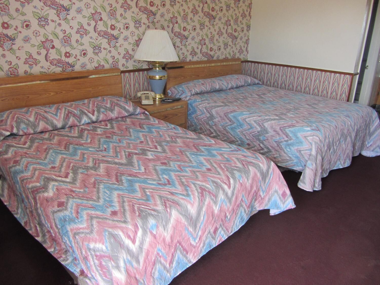 A room with two beds, each with a zigzag patterned bedspread, a nightstand with a lamp, and a floral wallpaper.
