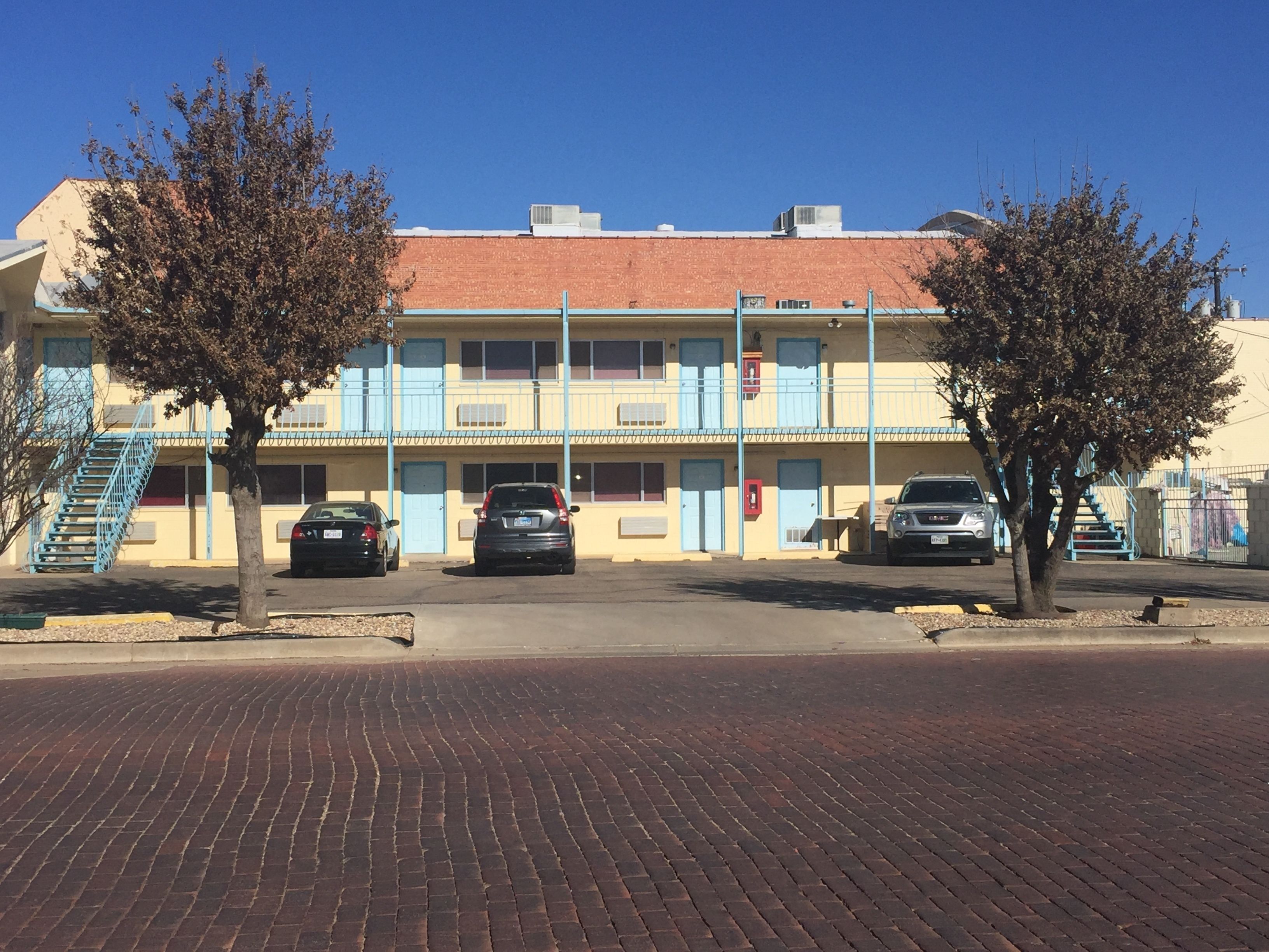 Un motel de dos pisos con techo de tejas rojas, ventanas con marcos azules y un estacionamiento con tres coches.