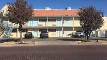 Un motel de dos pisos con techo de tejas rojas, ventanas con marcos azules y un estacionamiento con tres coches.