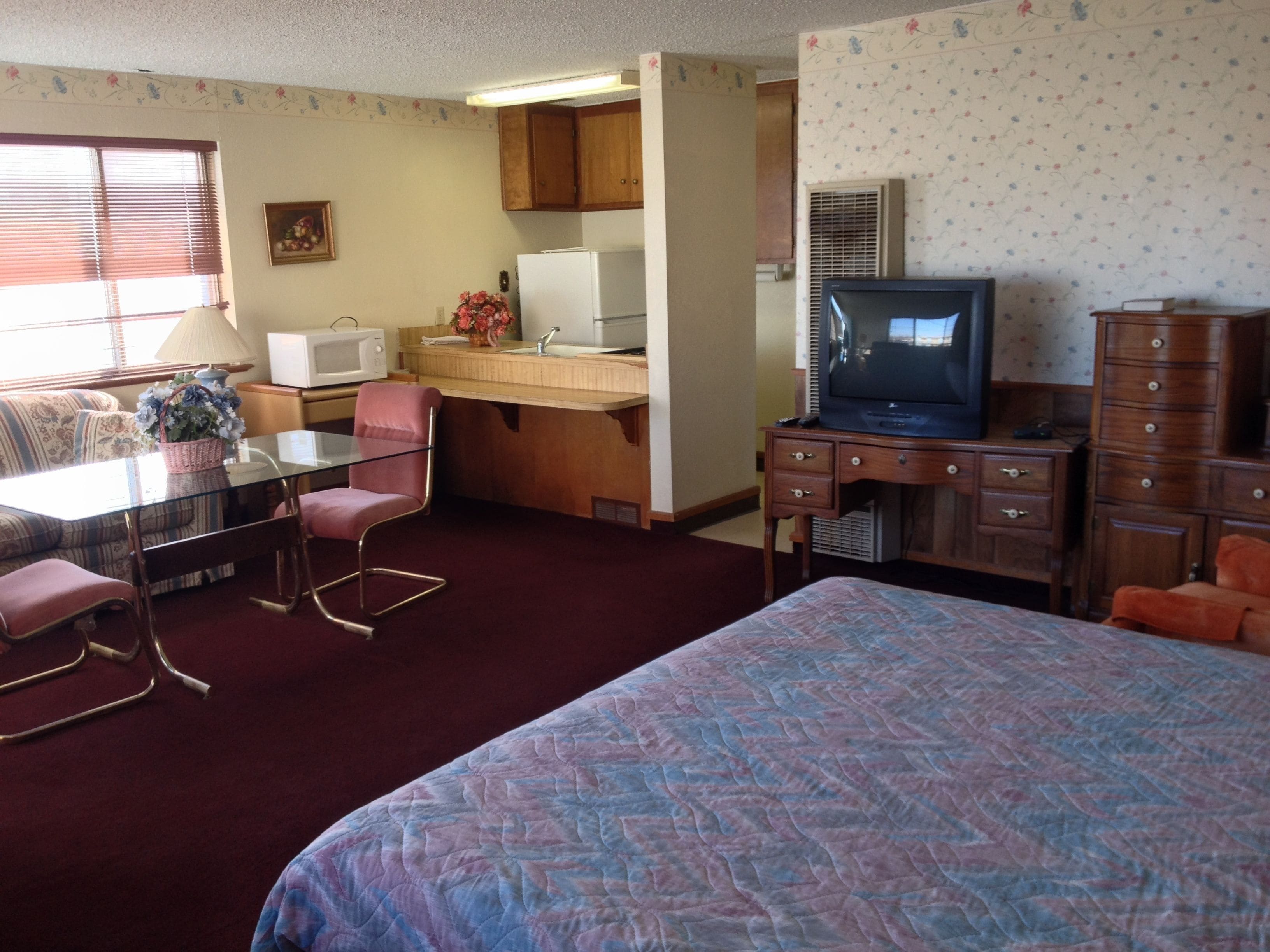 A compact hotel room with a bed, a desk with a chair, a kitchenette, a television, and a dresser.