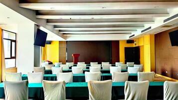A conference room with rows of tables and chairs, a flat-screen TV mounted on the wall, and a stage area.