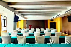Un salón de conferencias con filas de mesas y sillas, un televisor de pantalla plana montado en la pared y una zona de escenario.