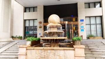 A fountain with a spherical top and multiple tiers in front of a building with a glass entrance.