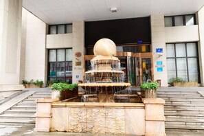 Ein Brunnen mit einer kugelförmigen Spitze und mehreren Stufen vor einem Gebäude mit Glastür.