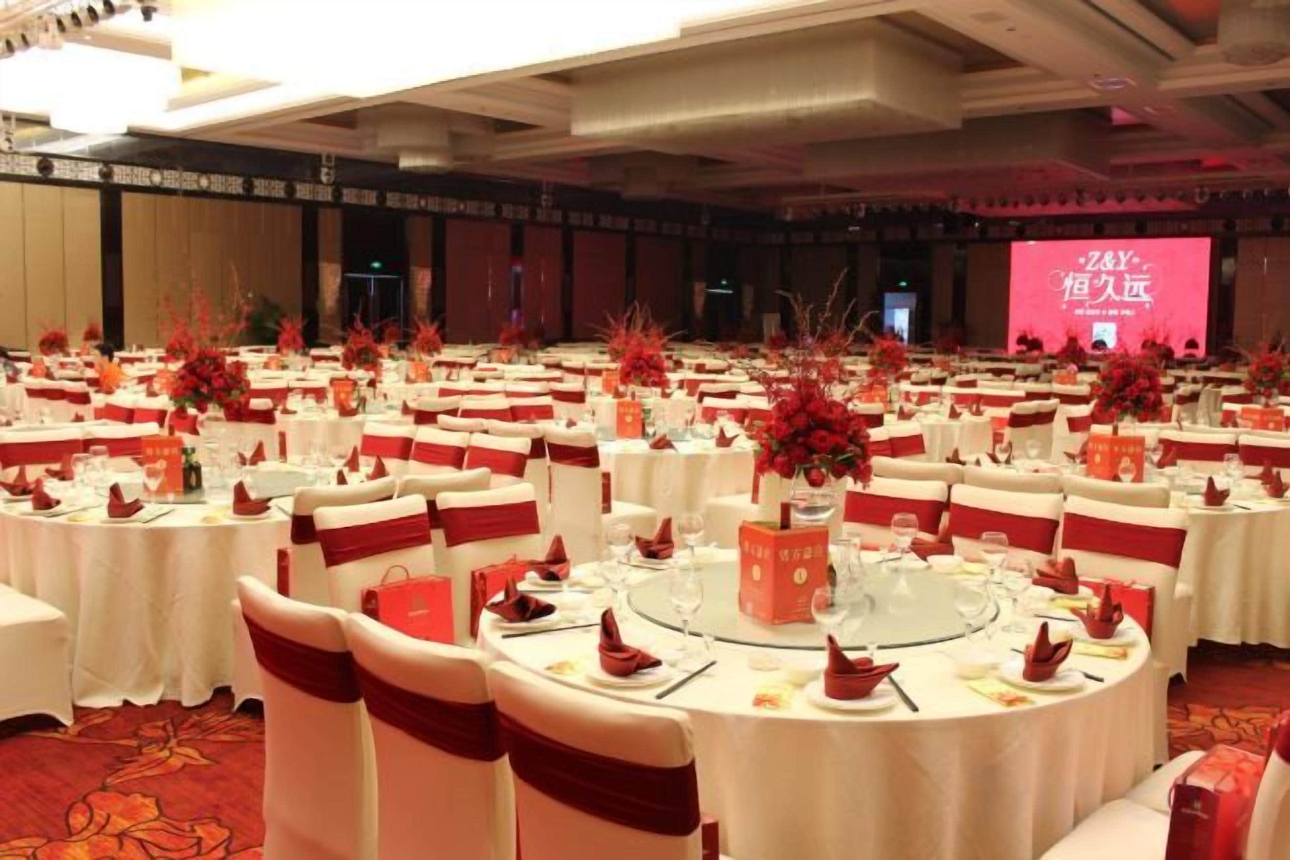 A banquet hall with round tables, red and white decor, and a stage with a pink screen.
